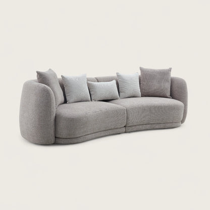 Divano dal design moderno e elegante in tessuto tortora con forma curva e asimmetrica, imbottitura avvolgente e design moderno per soggiorno raffinato.