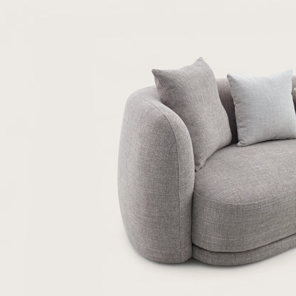 Divano dal design moderno e elegante in tessuto tortora con forma curva e asimmetrica, divano imbottito e braccioli avvolgente e confortevoli con angoli arrotondati.